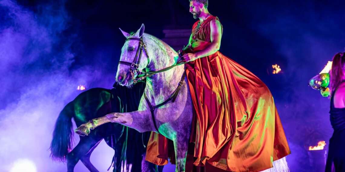 Un cavaliere in costume tipo akama rosso su un cavallo grigio che fa il passo spagnolo, durante uno spettacolo equestre illuminato da luci viola e verdi, e un fumo leggere sullo sfondo