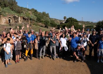 Bambini e adulti salutano con le mani alzate intorno a due asini, sullo sfondo di edifici in pietra e vegetazione a Pantelleria