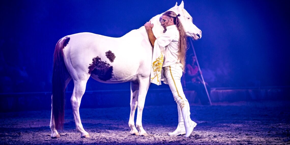 Rudj Bellini, in costume bianco e oro, abbraccia il collo del Quarab Mr. Martin nell’arena di Cavalluna illuminata di blu.