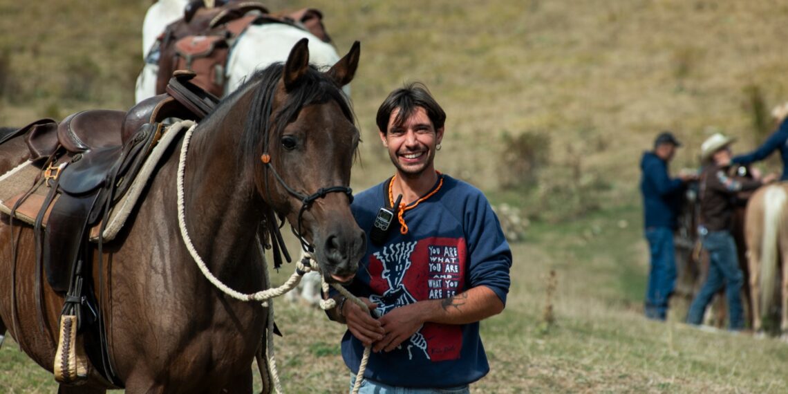Cavallo baio sellato e attrezzato per il trekking, accanto a un cavaliere sorridente durante una sosta all'aperto.