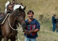 Cavallo baio sellato e attrezzato per il trekking, accanto a un cavaliere sorridente durante una sosta all'aperto.