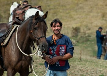 Cavallo baio sellato e attrezzato per il trekking, accanto a un cavaliere sorridente durante una sosta all'aperto.