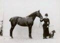 Fotografia storica del 1890 circa: una donna in abito da amazzone ottocentesco, con cappello a bombetta, tiene per le redini un cavallo scuro. Accanto a lei un piccolo barboncino.