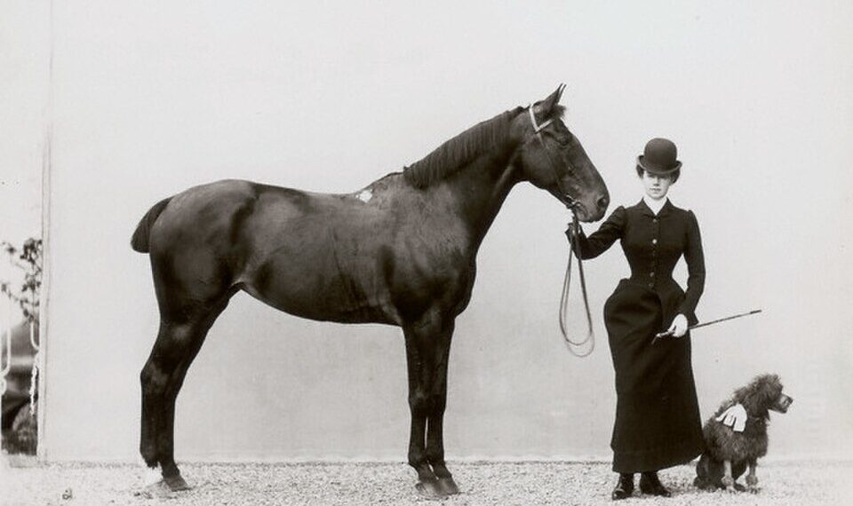 Fotografia storica del 1890 circa: una donna in abito da amazzone ottocentesco, con cappello a bombetta, tiene per le redini un cavallo scuro. Accanto a lei un piccolo barboncino.