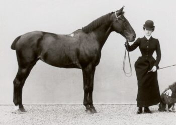 Fotografia storica del 1890 circa: una donna in abito da amazzone ottocentesco, con cappello a bombetta, tiene per le redini un cavallo scuro. Accanto a lei un piccolo barboncino.