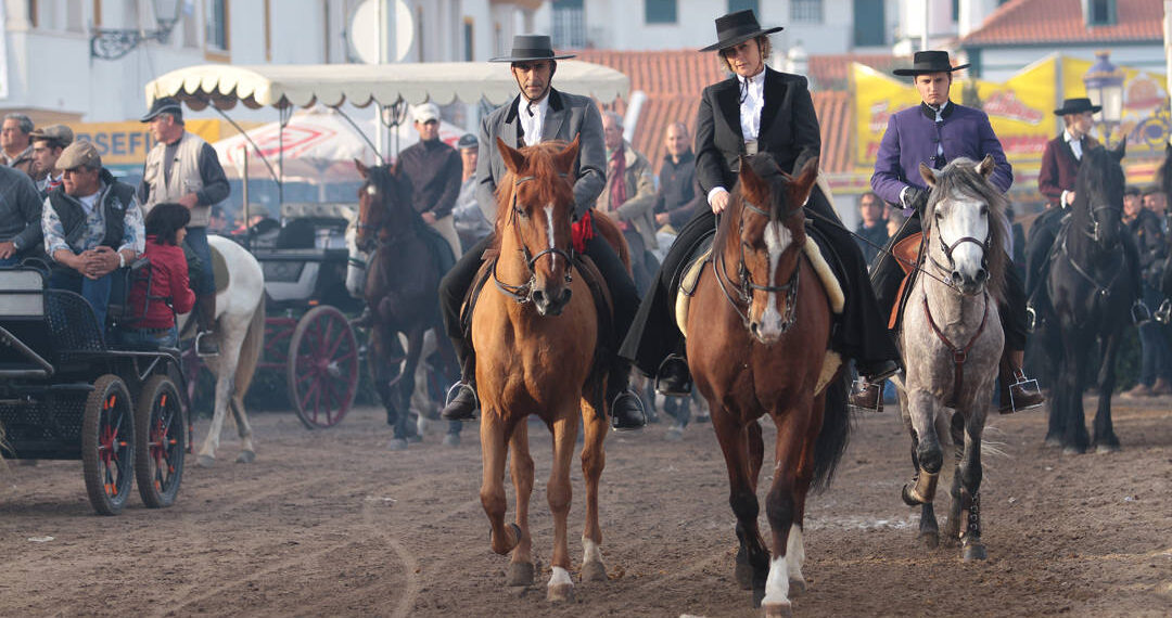 Tre persone a cavallo in abiti tradizionali portoghesi attraversano la Fiera di Golegã, con carrozze, altri cavalieri e pubblico sullo sfondo