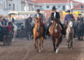 Tre persone a cavallo in abiti tradizionali portoghesi attraversano la Fiera di Golegã, con carrozze, altri cavalieri e pubblico sullo sfondo