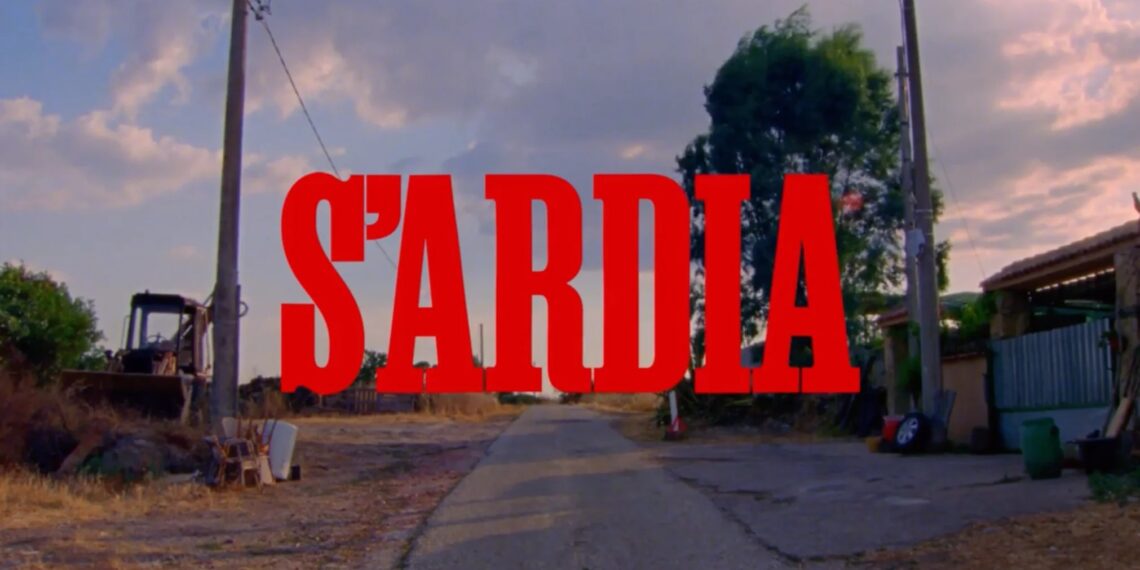 Titolo rosso «S’Ardia» sovrapposto a una strada rurale, fra edifici agricoli e alberi, sotto un cielo con nuvole rosate