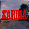 Titolo rosso «S’Ardia» sovrapposto a una strada rurale, fra edifici agricoli e alberi, sotto un cielo con nuvole rosate