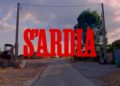 Titolo rosso «S’Ardia» sovrapposto a una strada rurale, fra edifici agricoli e alberi, sotto un cielo con nuvole rosate